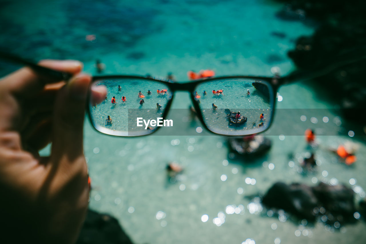 Cropped hand holding eyeglasses against people in sea