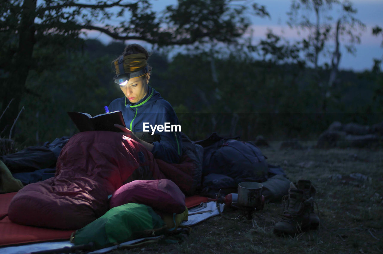 Woman writing on book while camping on field at dusk