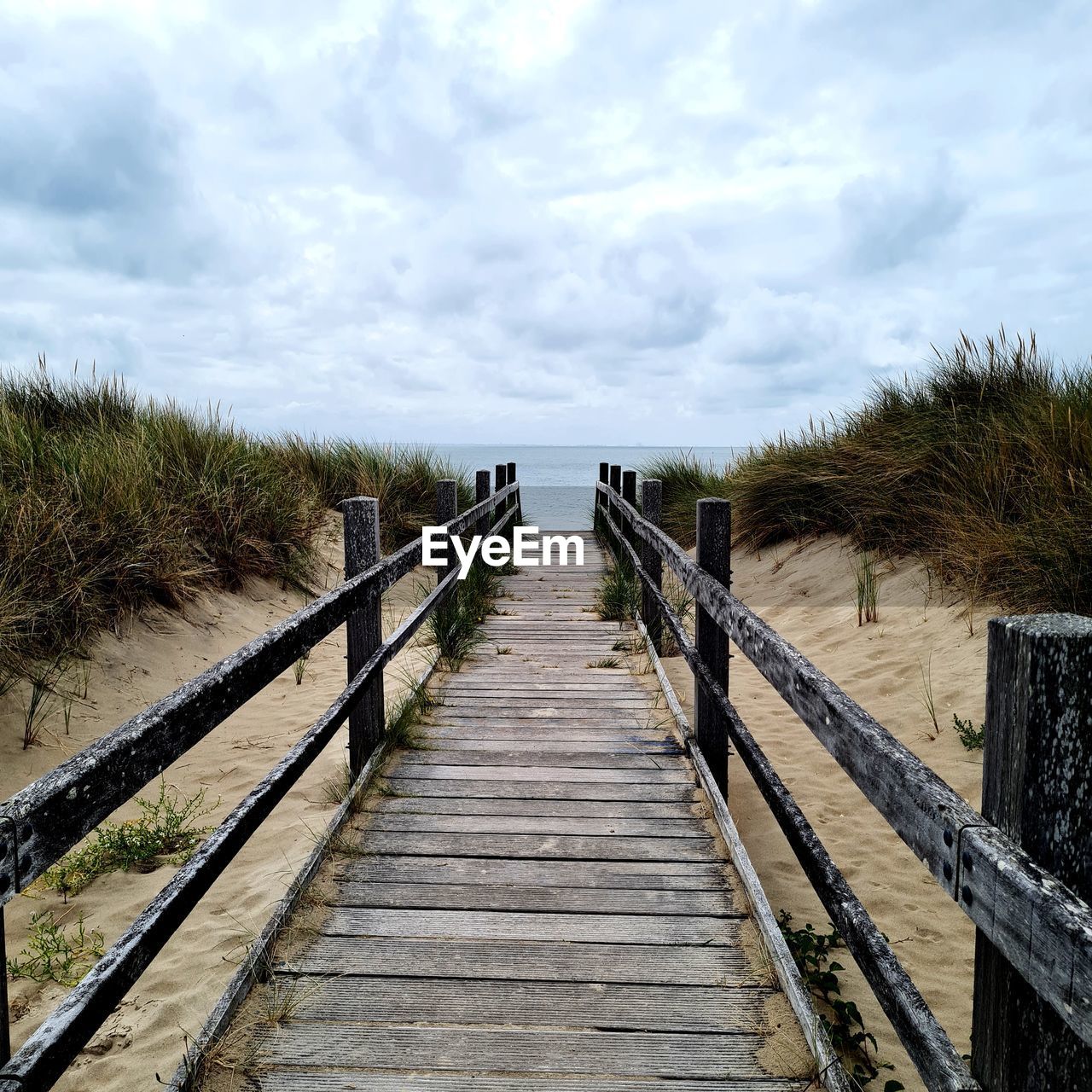 walkway, sky, the way forward, cloud, boardwalk, nature, wood, land, water, footpath, railing, plant, tranquility, beach, scenics - nature, no people, coast, diminishing perspective, sea, day, tranquil scene, beauty in nature, landscape, non-urban scene, architecture, footbridge, bridge, outdoors, environment, built structure, tree, horizon, waterway, pier, grass, shore, vanishing point, track, travel destinations, sand