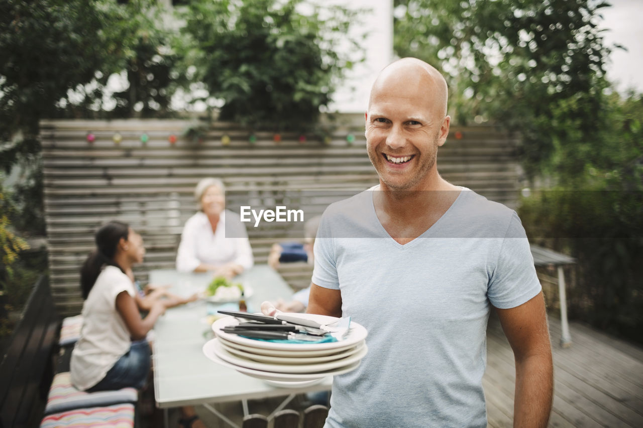 Portrait of happy man holding plates with family sitting at outdoor table in yard