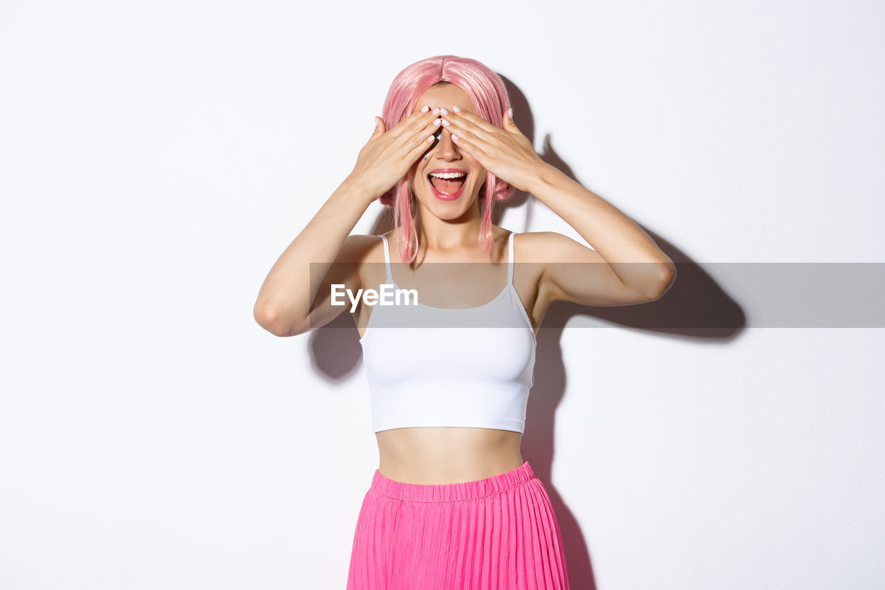 Young smiling woman covering eyes standing against wall