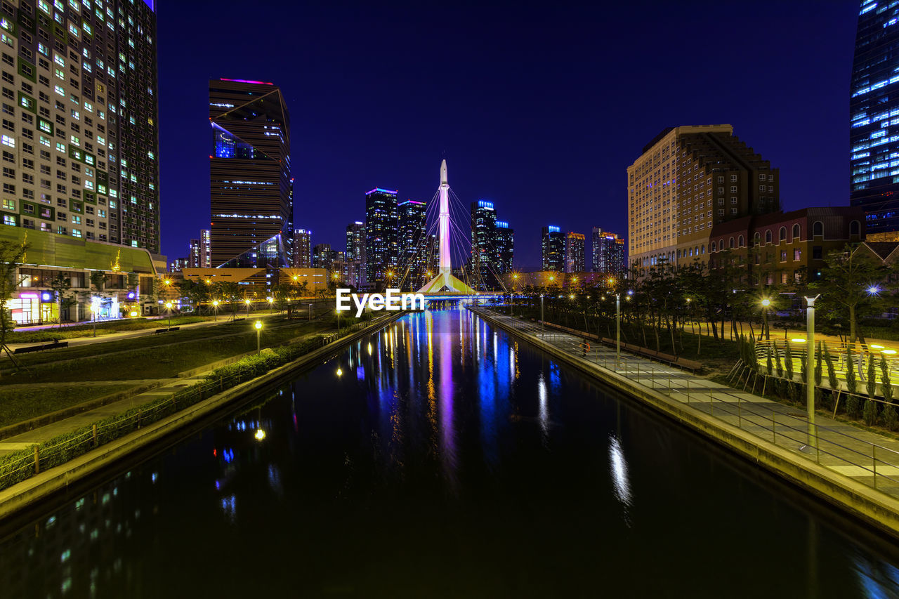 Illuminated modern buildings by river against sky at night