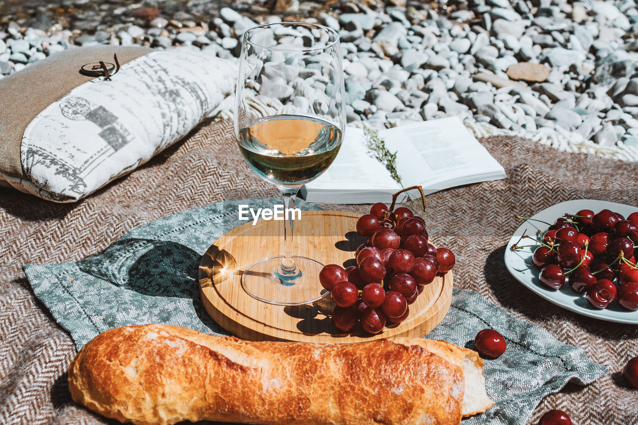 Summer romantic rustic picnic on the beach, white wine glass, baguette, cherries, grape, open book