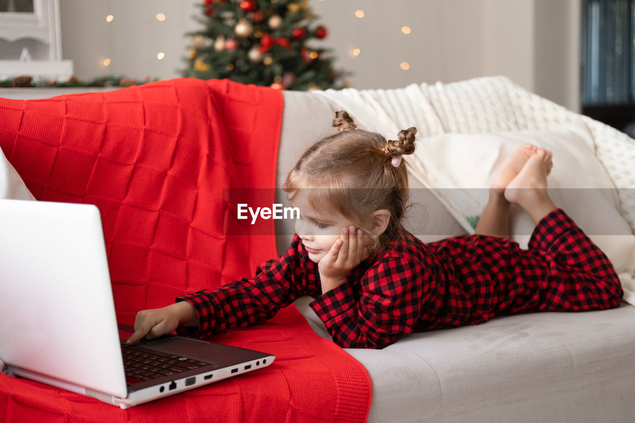 Cute little girl in red christmas pajama using laptop, having video chat, lying on couch