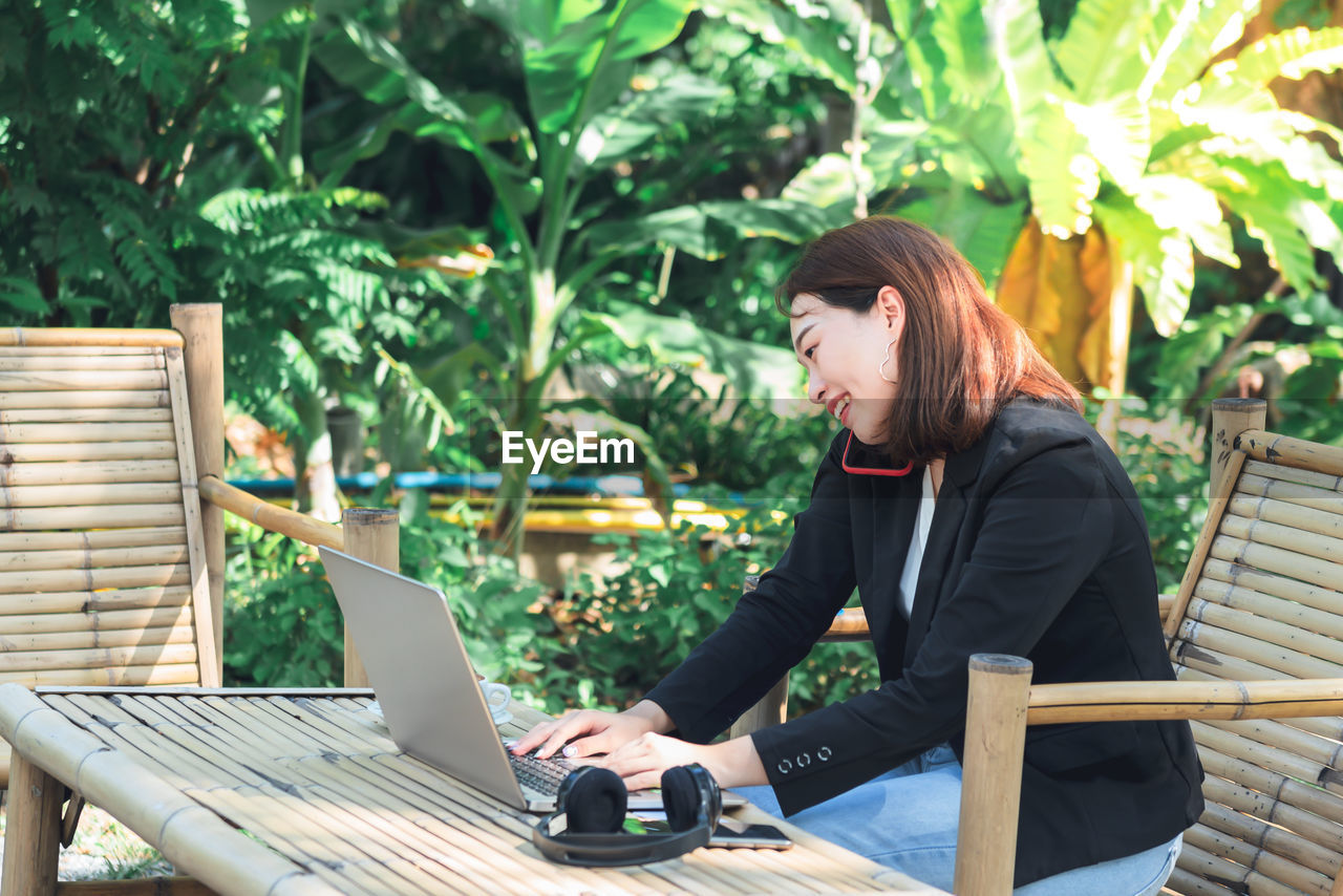 YOUNG WOMAN LOOKING AT CAMERA WHILE SITTING ON TABLE AGAINST PLANTS