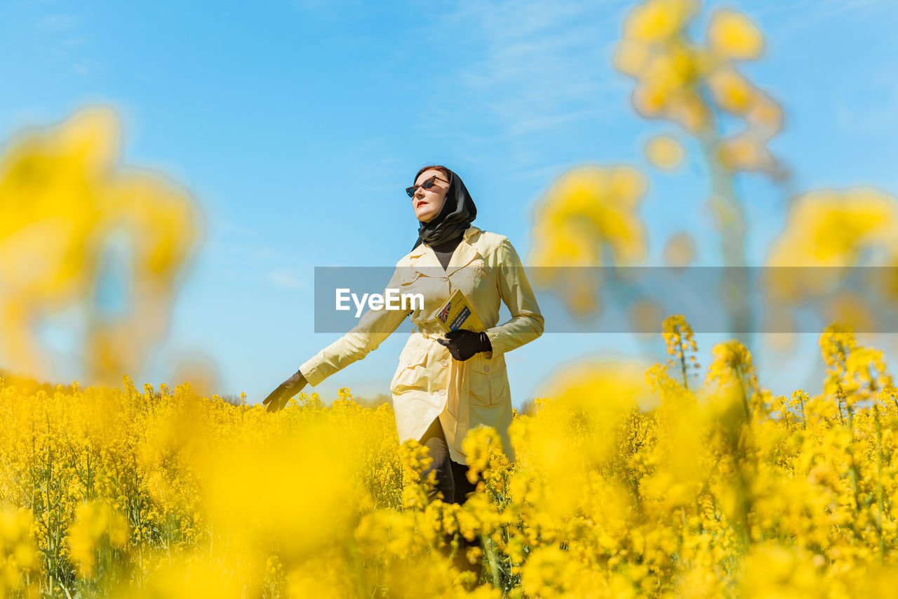 A young stylish woman poses among a blooming yellow field of rapeseed under a bright spring sky