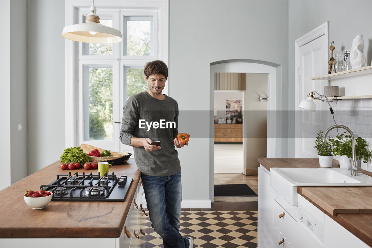 Man using smartphone and holding bell pepper in kitchen