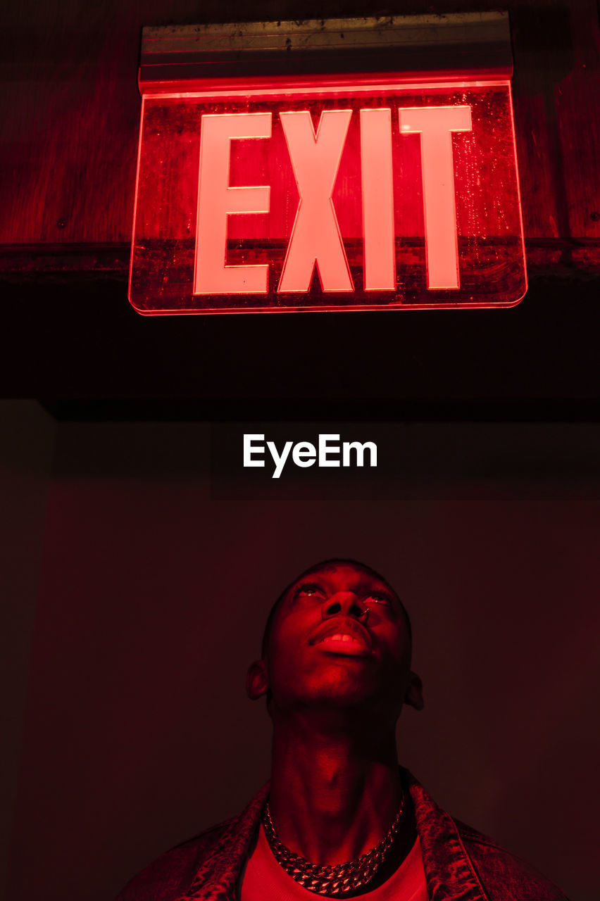 From below crop african american young male looking up on illuminated tablet exit above head in red dark light