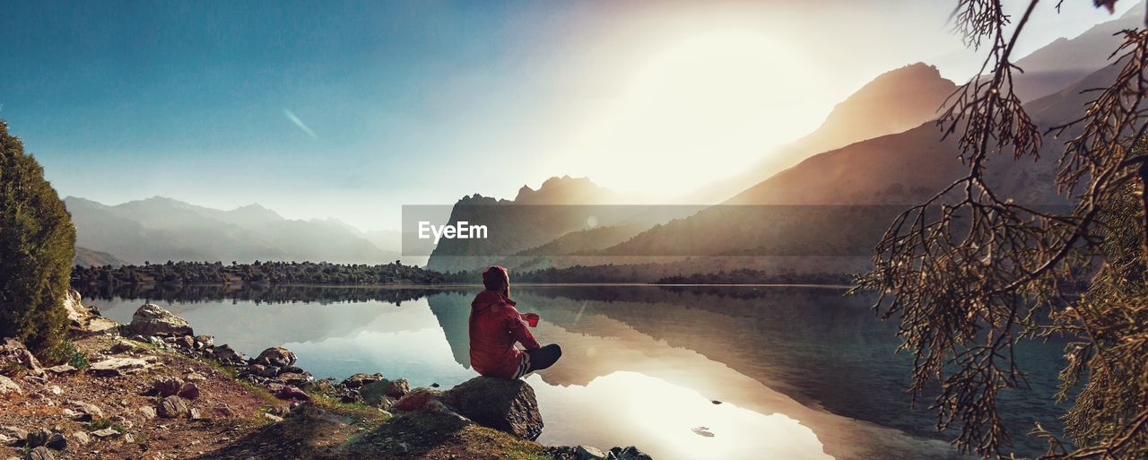 Man looking at view while sitting on rock by lake against mountains and sky during sunset