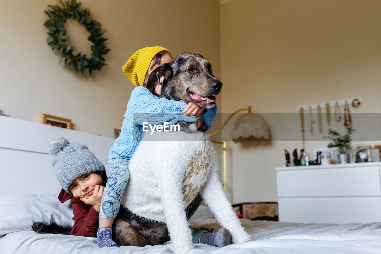 Two children wearing warm hats hug their dog which is wearing sweater