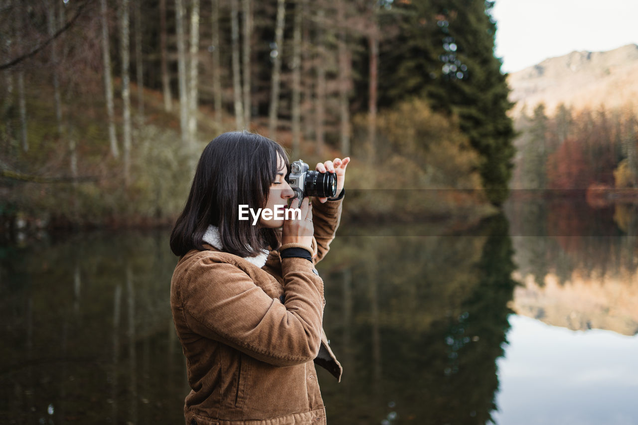 Side view of young female traveler in brown jacket taking picture with photo camera while standing next to lake surrounded with forest in autumn day
