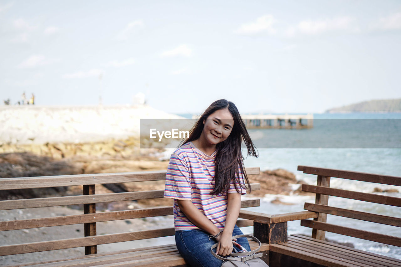 Portrait of smiling young woman sitting on bench by sea