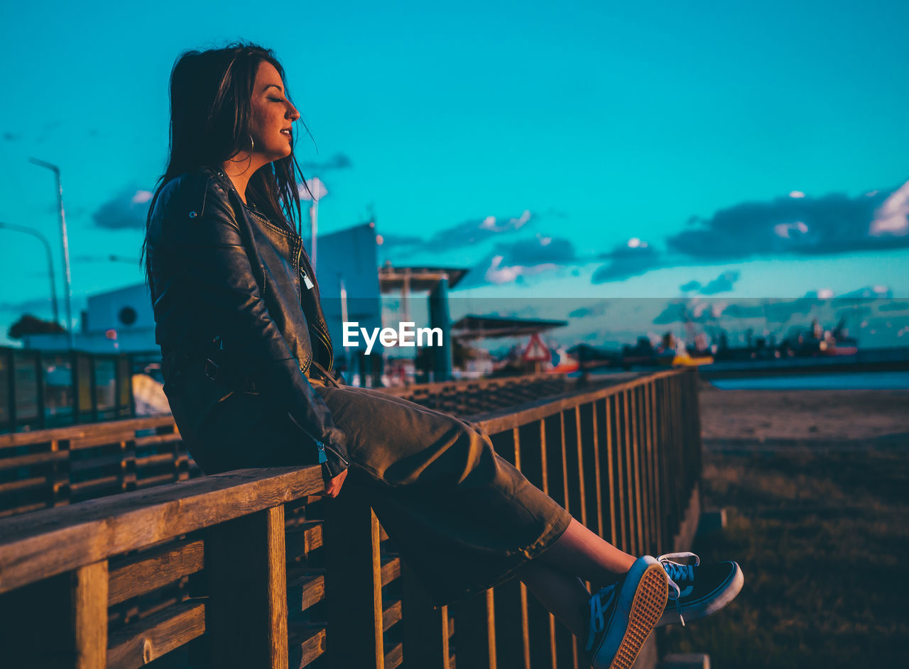 Young woman sitting on railing at beach against sky