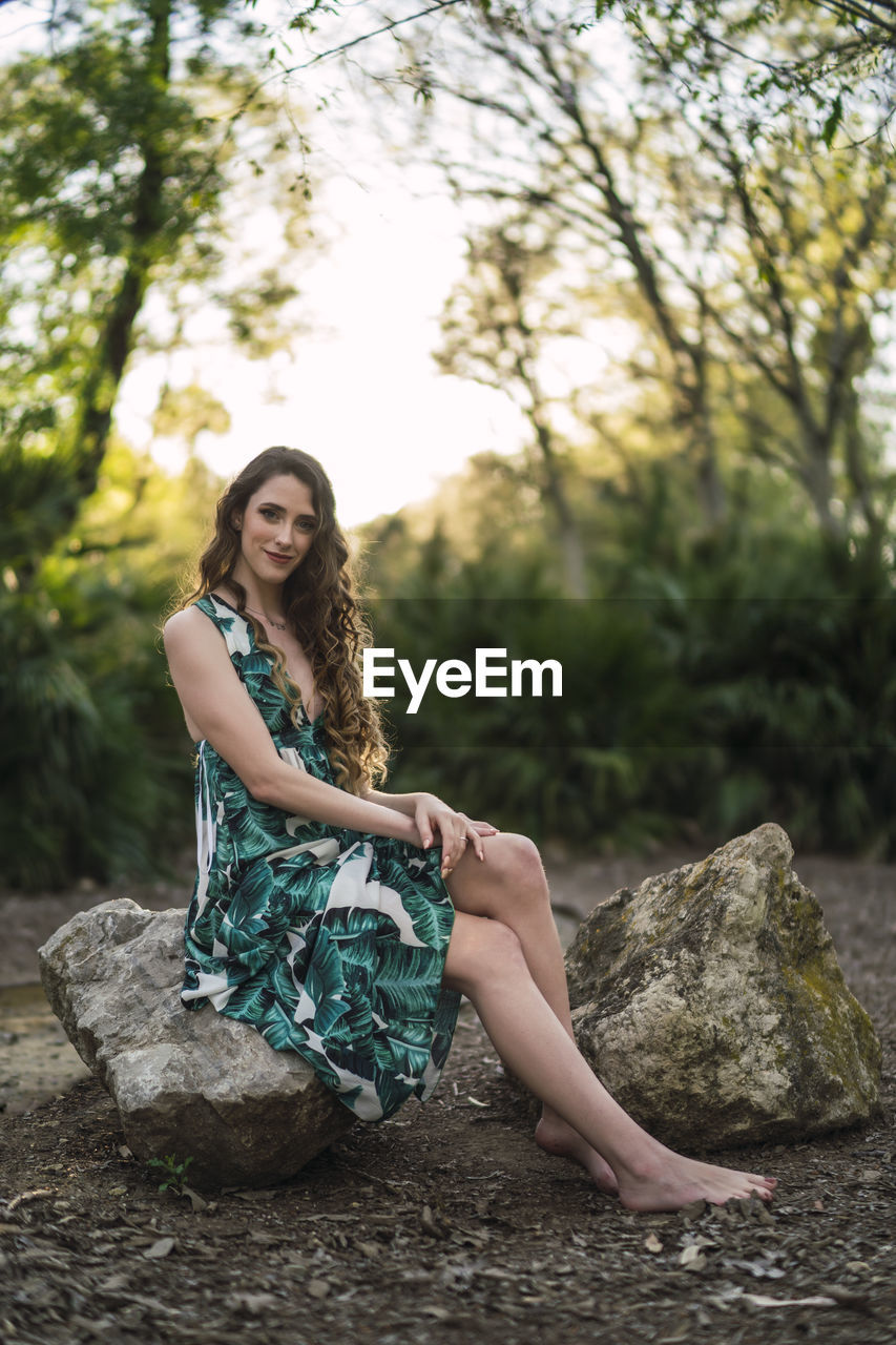 Full body content young female wearing trendy maxi sundress and looking at camera with smile while sitting on big stone in verdant sunny woodland