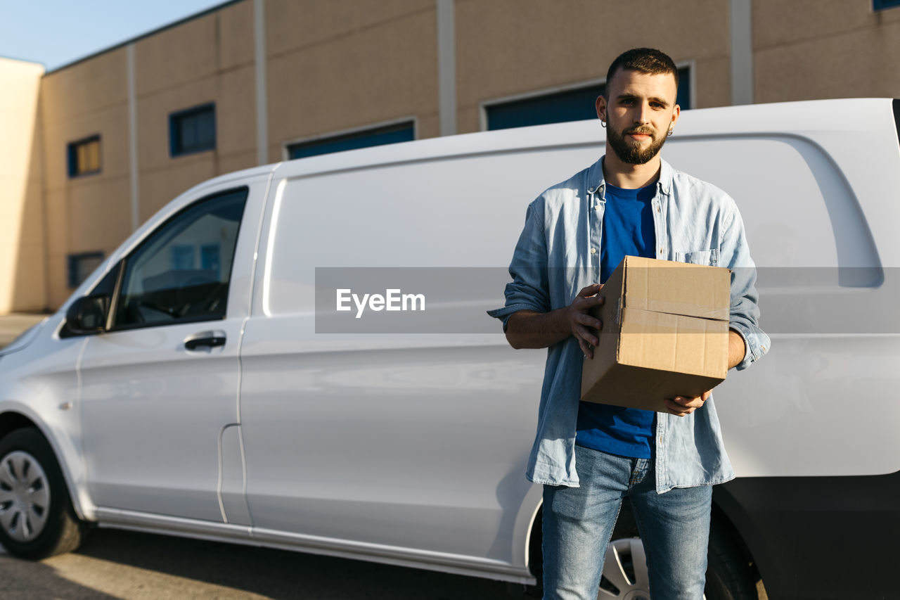 Young delivery man with package standing in front of van