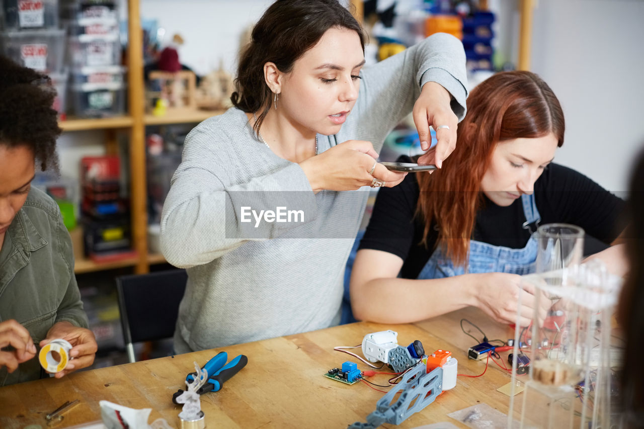 Young female inventor photographing model on table while sitting amidst colleagues at workshop