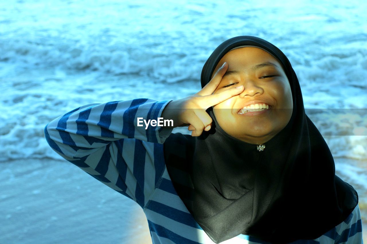 Portrait of cheerful teenage girl gesturing peace sign at beach