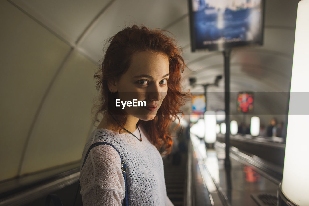 High angle side view of young female passenger in casual outfit with red curly hair looking at camera with smile while going down subway escalator in saint petersburg