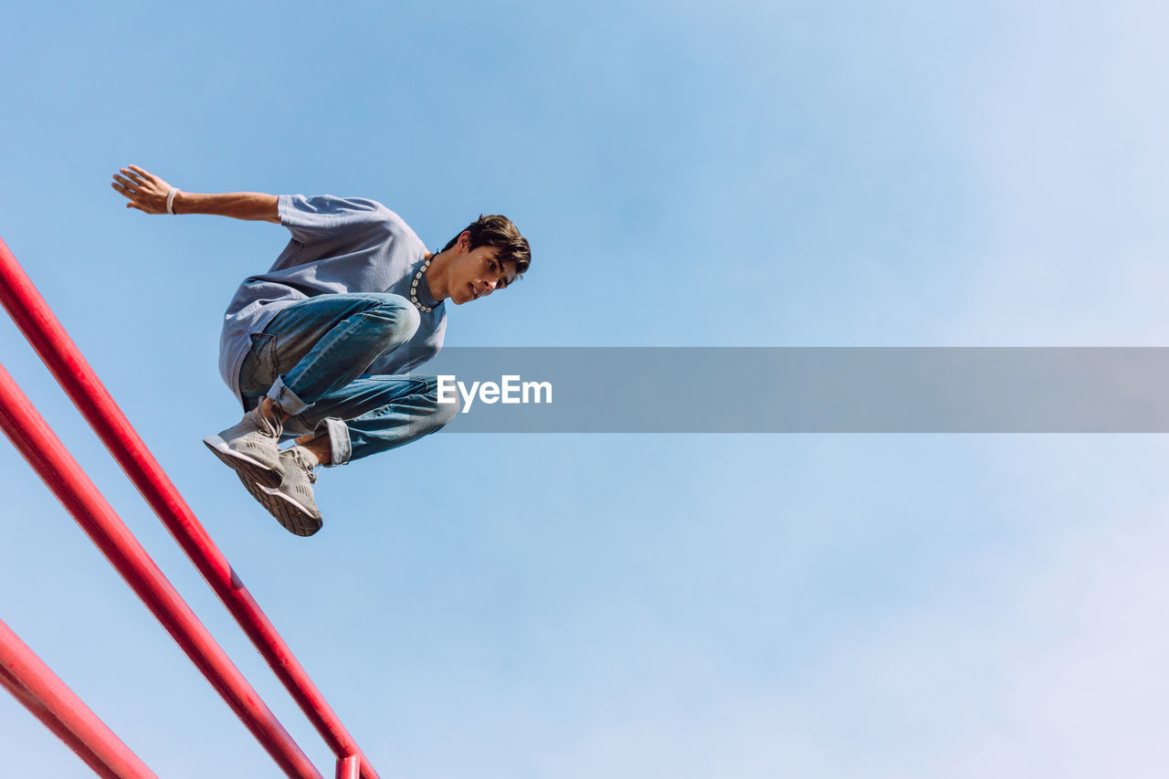 From below of brave male jumping over metal fence in street and showing parkour trick against blue sky