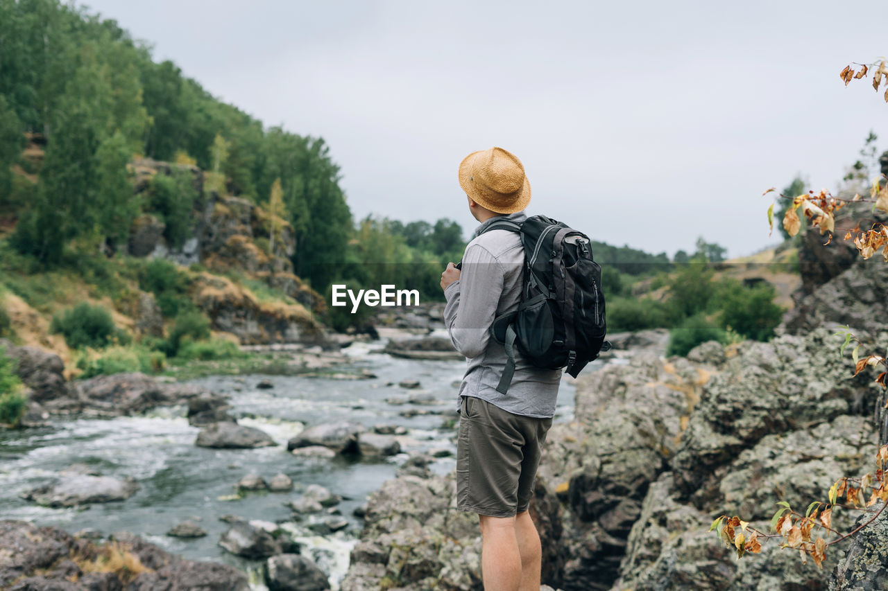 Young man traveler in straw hat with backpack at mountain river, local travel 