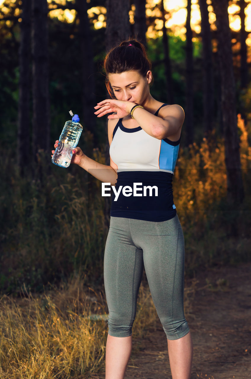 Young woman drinking water while standing against trees in forest during sunset