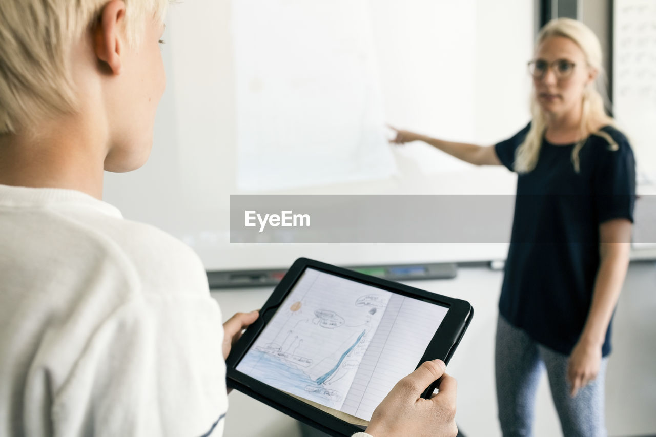 Cropped image of boy holding digital tablet with teacher explaining in classroom