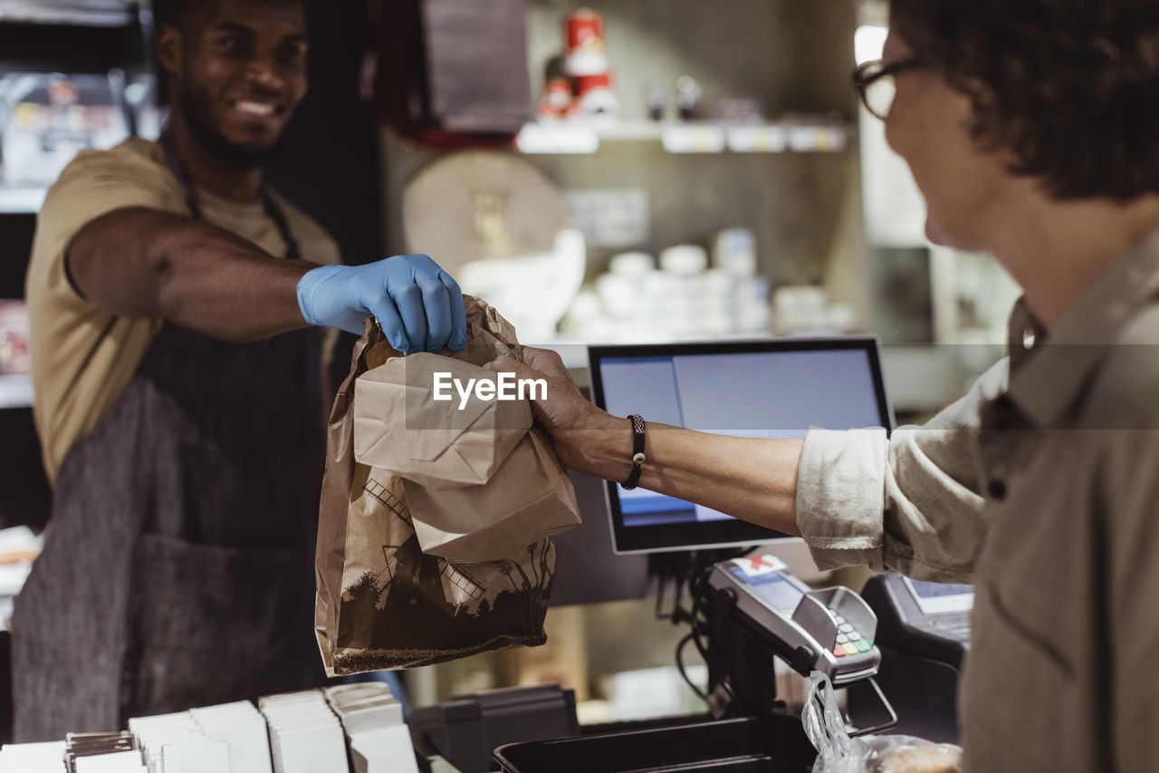 Male owner giving package order to female customer in delicatessen shop