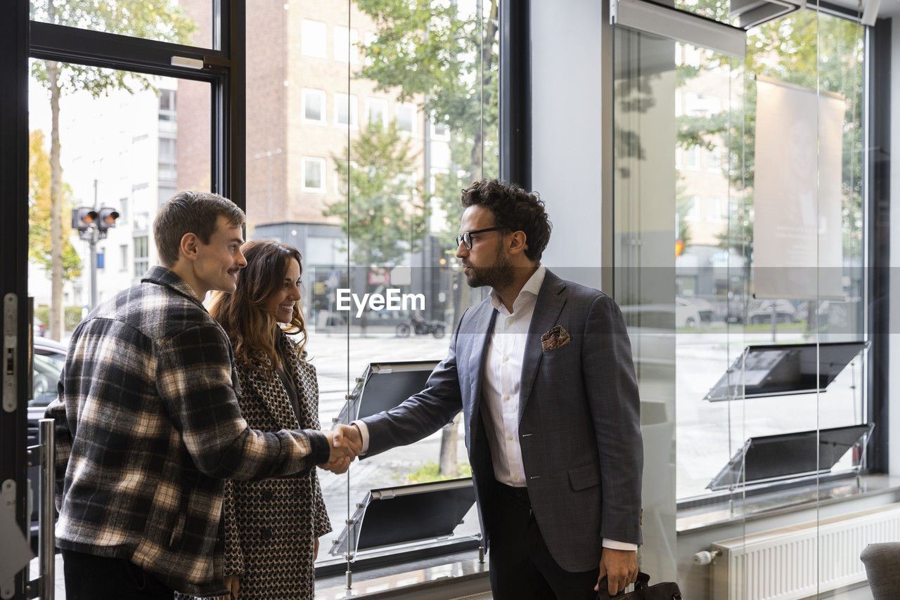 Happy couple shaking hand with real estate agent at entrance of office