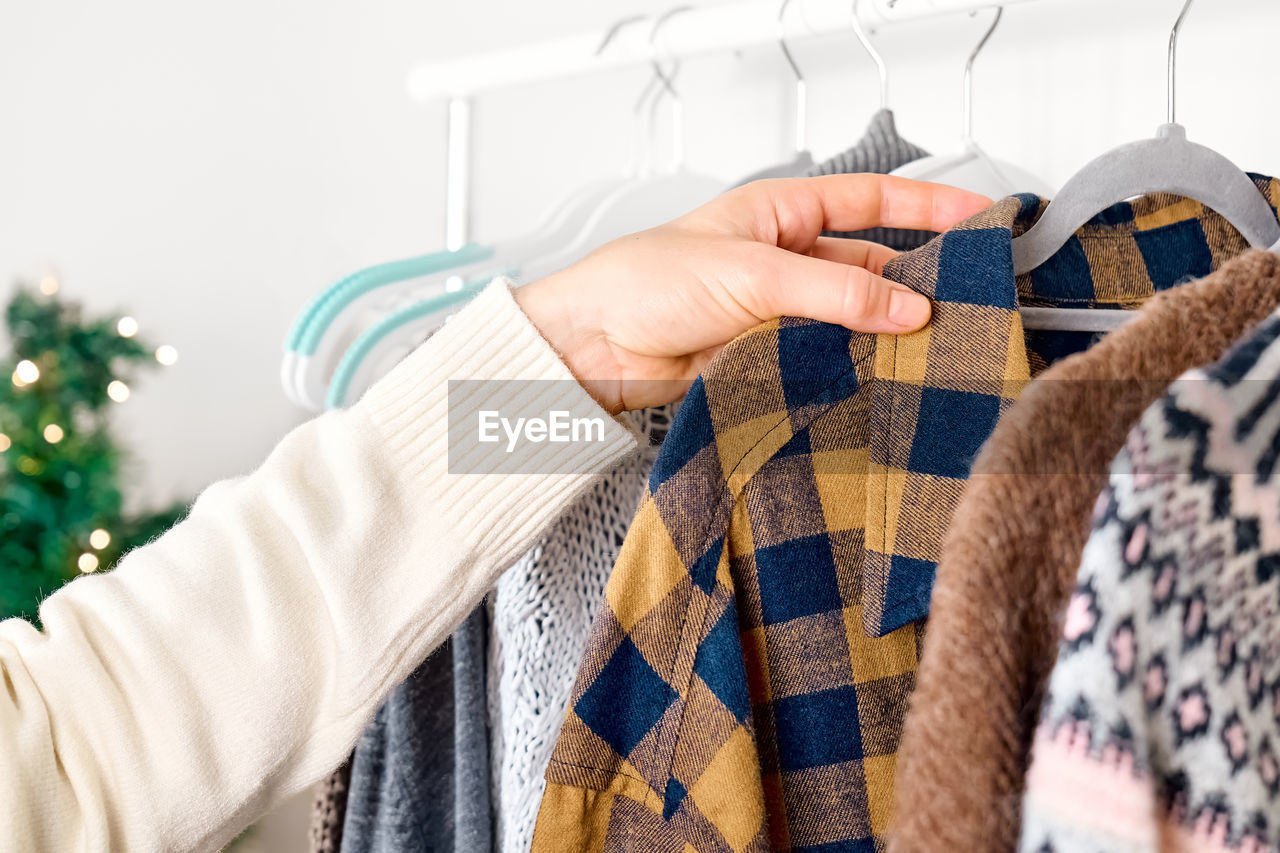 Woman's hand taking knitted sweater on hanger on clothes rack in shop with blurred christmas tree