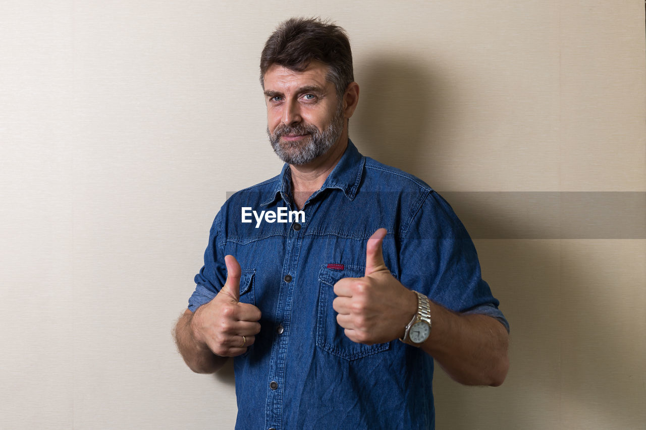 Portrait of mature man gesturing thumbs up while standing against wall