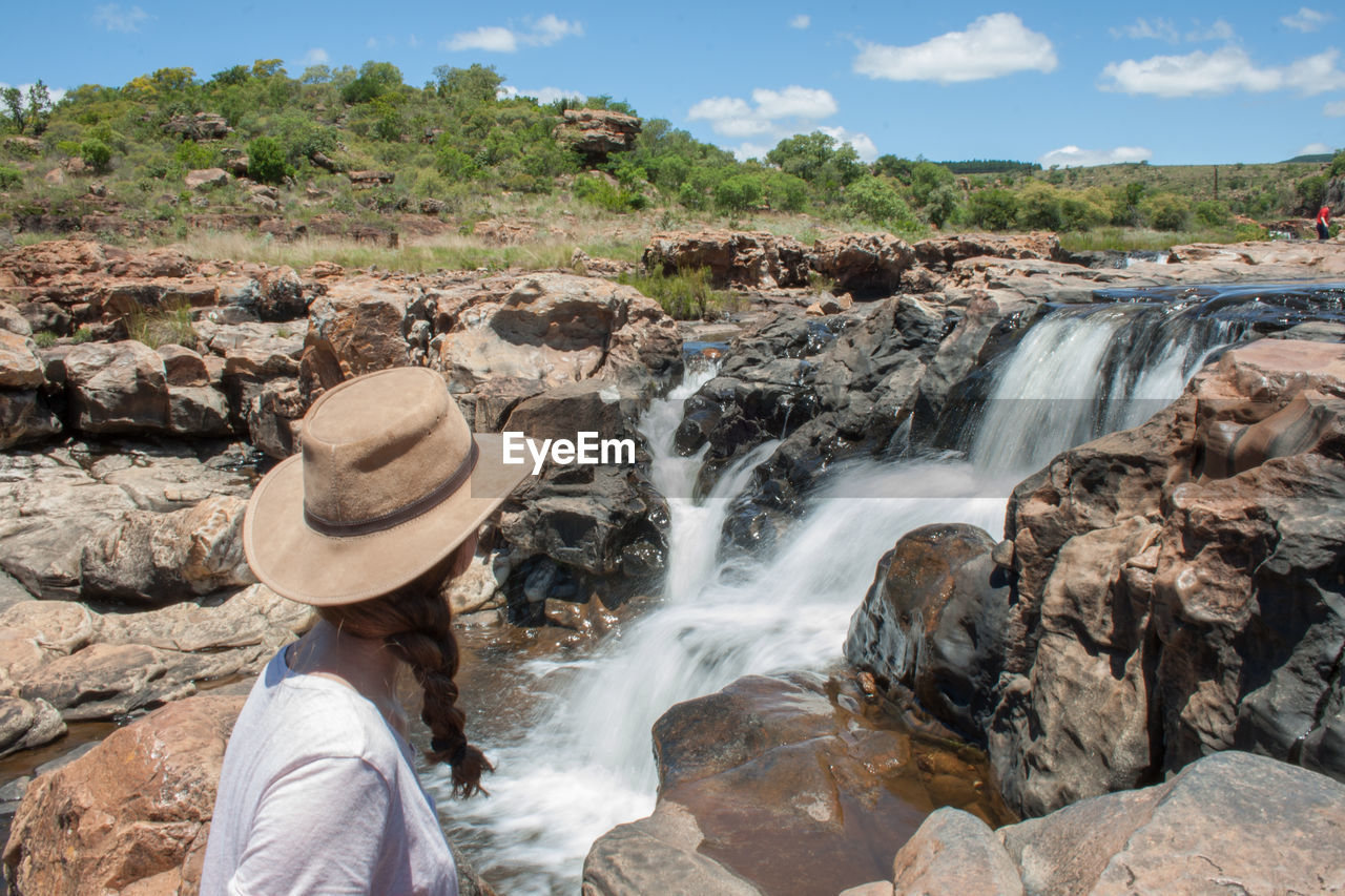 MAN STANDING ON ROCKS AGAINST WATERFALL