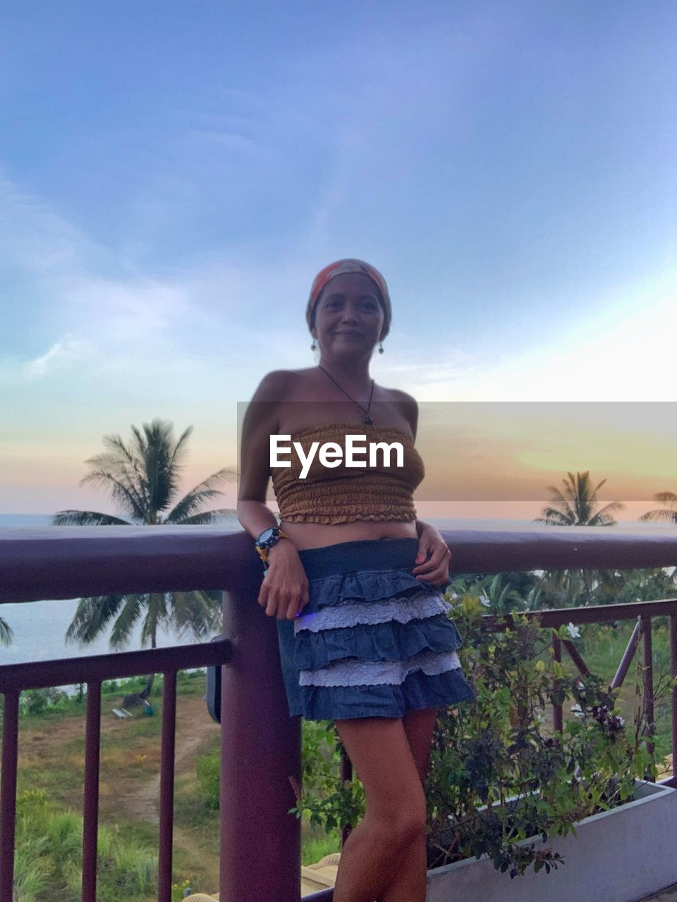 vacation, sky, one person, railing, nature, standing, blue, adult, casual clothing, young adult, lifestyles, leisure activity, women, clothing, sunset, water, shorts, architecture, full length, relaxation, cloud, three quarter length, front view, tropical climate, trip, holiday, outdoors, day, looking, sunlight, smiling, person, plant, happiness, beauty in nature, emotion, tree, tranquility, fashion, portrait, palm tree, travel, looking away, dress, travel destinations, sea
