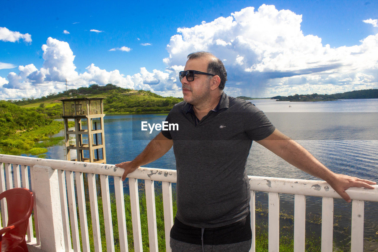 vacation, water, sky, one person, railing, adult, glasses, nature, cloud, sunglasses, men, standing, casual clothing, fashion, leisure activity, sea, day, holiday, front view, lifestyles, trip, architecture, mature adult, scenics - nature, clothing, three quarter length, outdoors, fence, sunlight, travel, person, beard, beauty in nature, summer, mountain, beach, relaxation, balcony, portrait, facial hair, deck, blue, wood, built structure, waist up, travel destinations