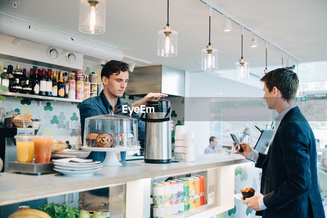 Businessman holding muffin and using phone while owner making coffee at office cafeteria