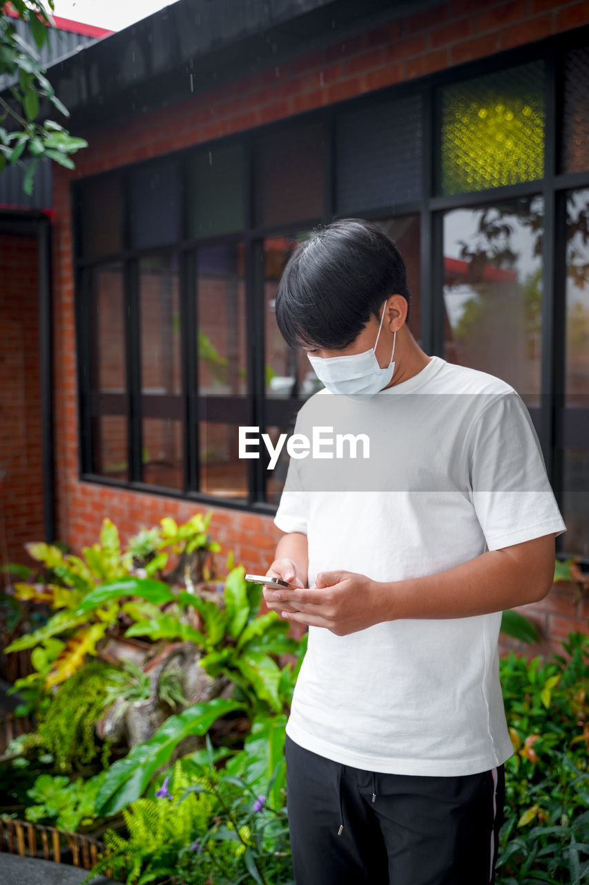 A handsome young teenage boy wear mask playing on smart mobile phone waiting out of cafeteria.