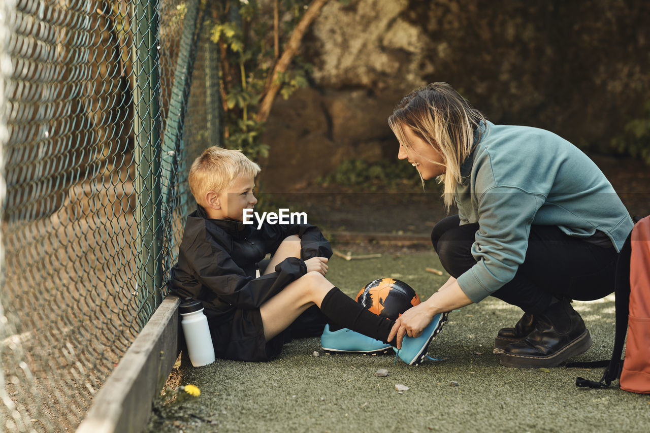 Happy woman tying shoelace of son sitting by fence in ground