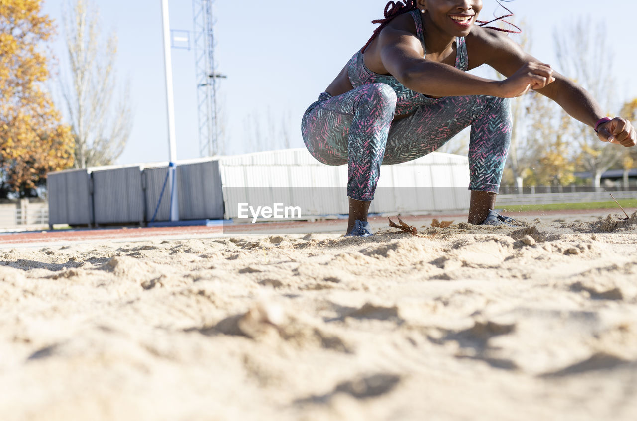 Smiling female athlete landing on sand in long jump during sunny day