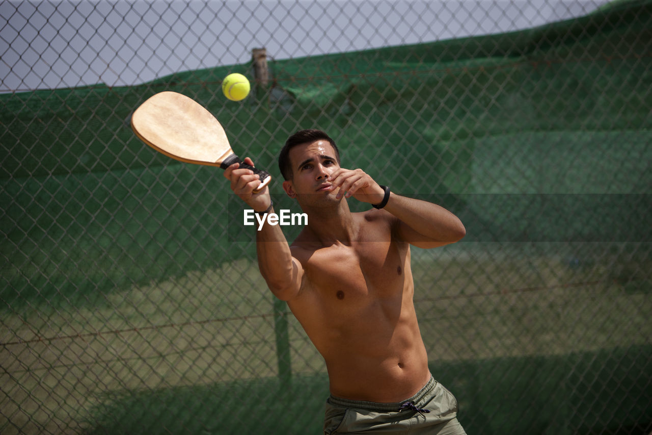 Shirtless young man playing tennis while standing against chainlink fence