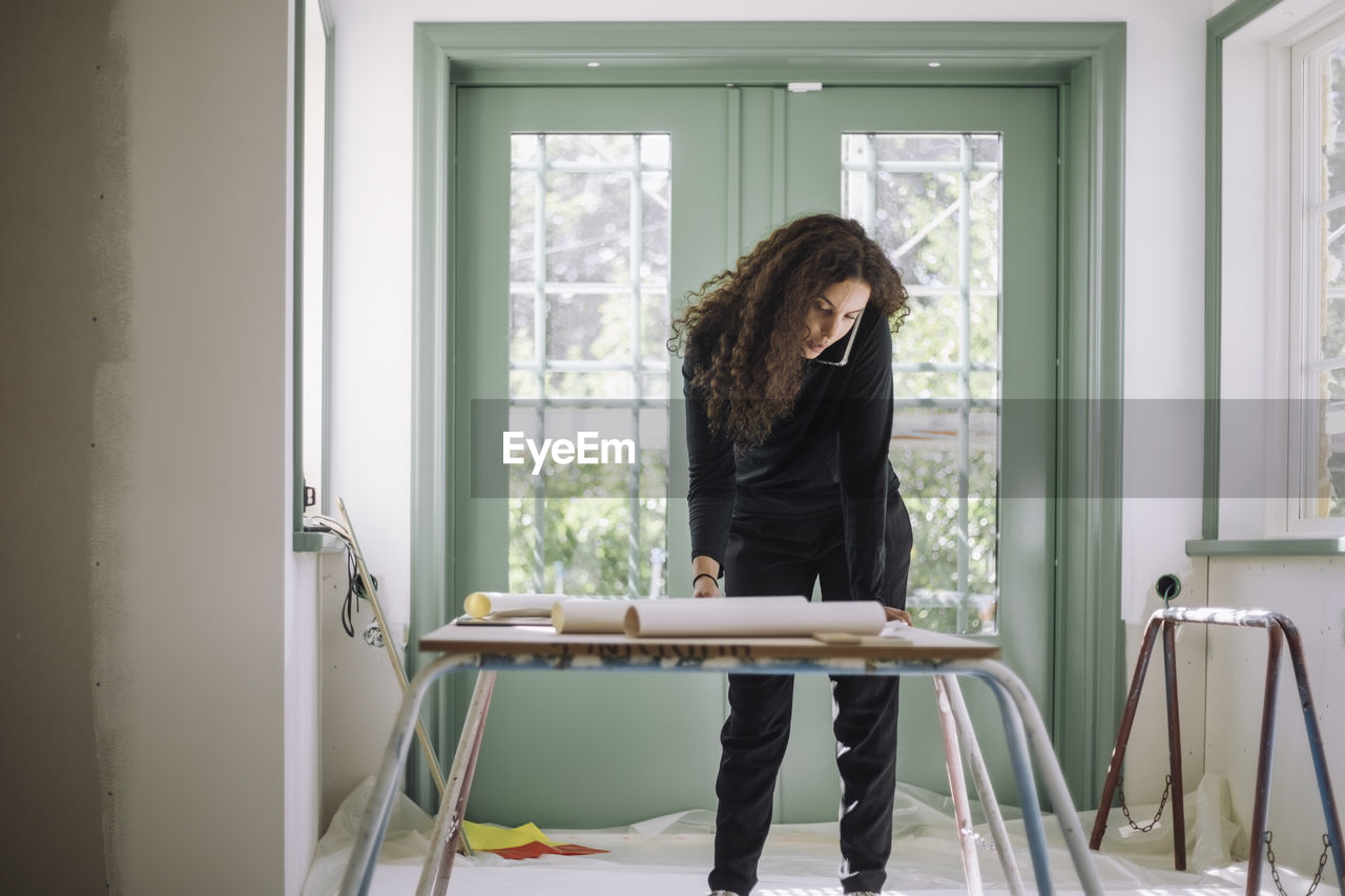 Female design professional talking on smart phone while examining blueprints on table at under construction apartment