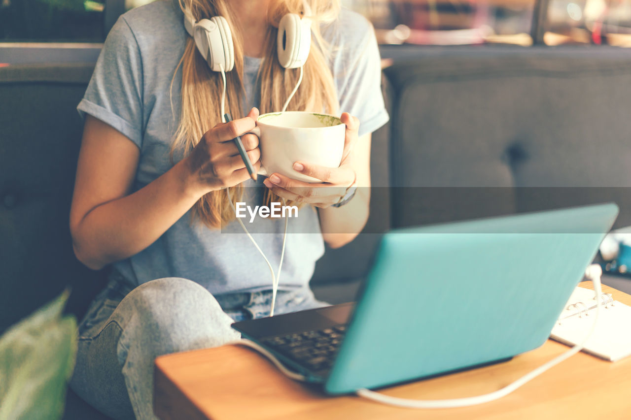  woman with headphones and a laptop at a table in a coffee shop holding a cup with a drink