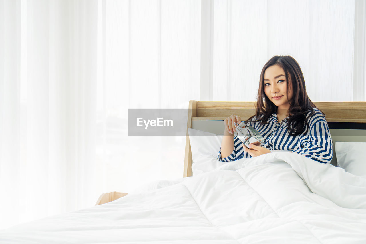 PORTRAIT OF BEAUTIFUL YOUNG WOMAN SITTING ON BED IN BEDROOM
