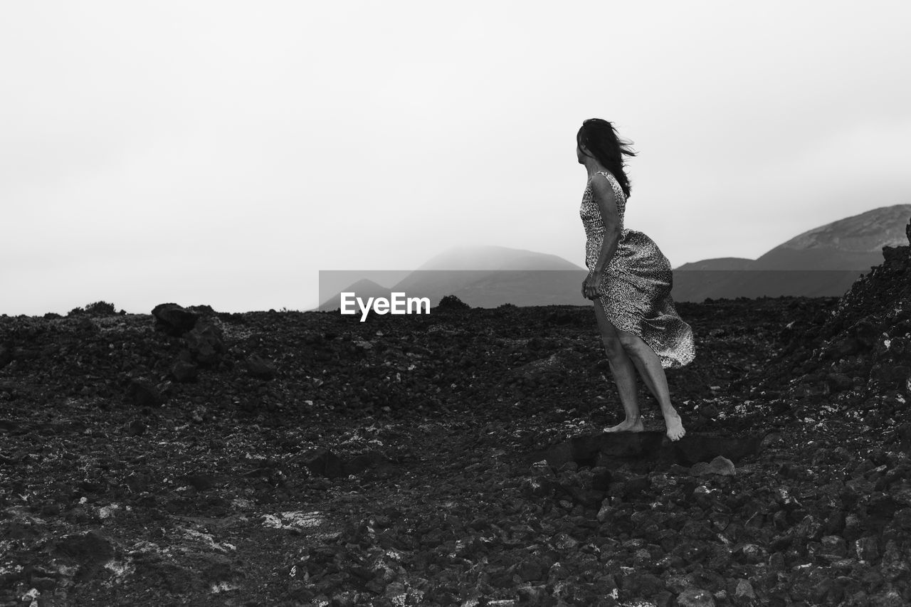 Side view of woman standing on volcanic rocks against sky