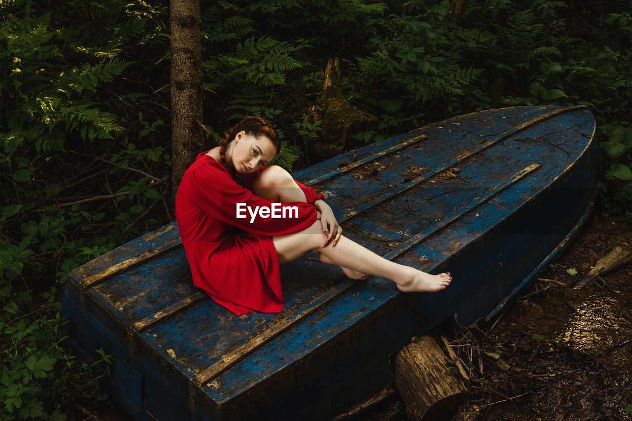 Young woman sitting on old boat against trees in forest