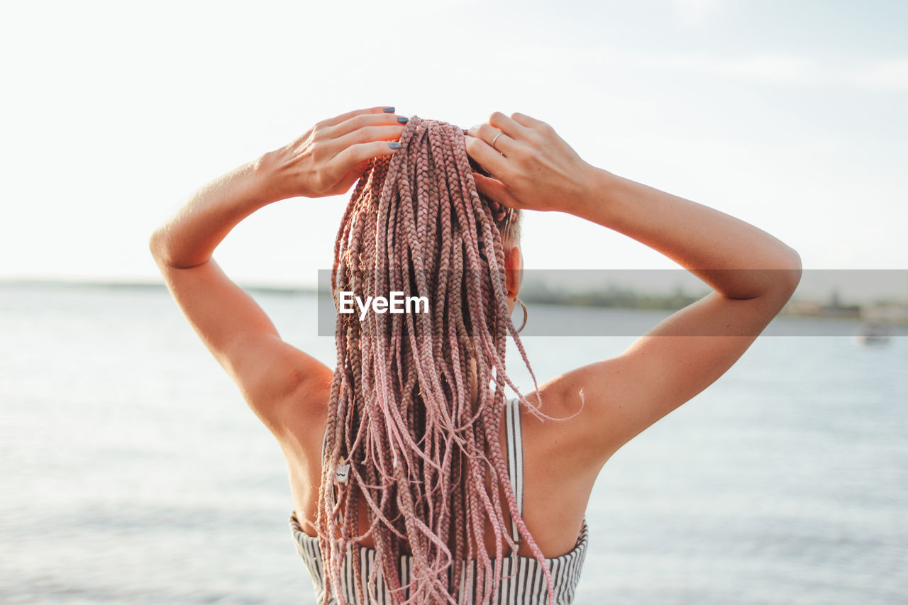 Rear view of woman with hand in hair standing at beach against sky