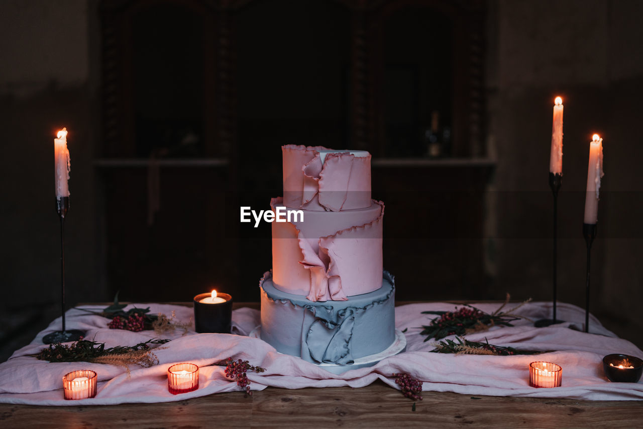 Delicious tiered cake served on wooden table and surrounded by burning candles in dark room