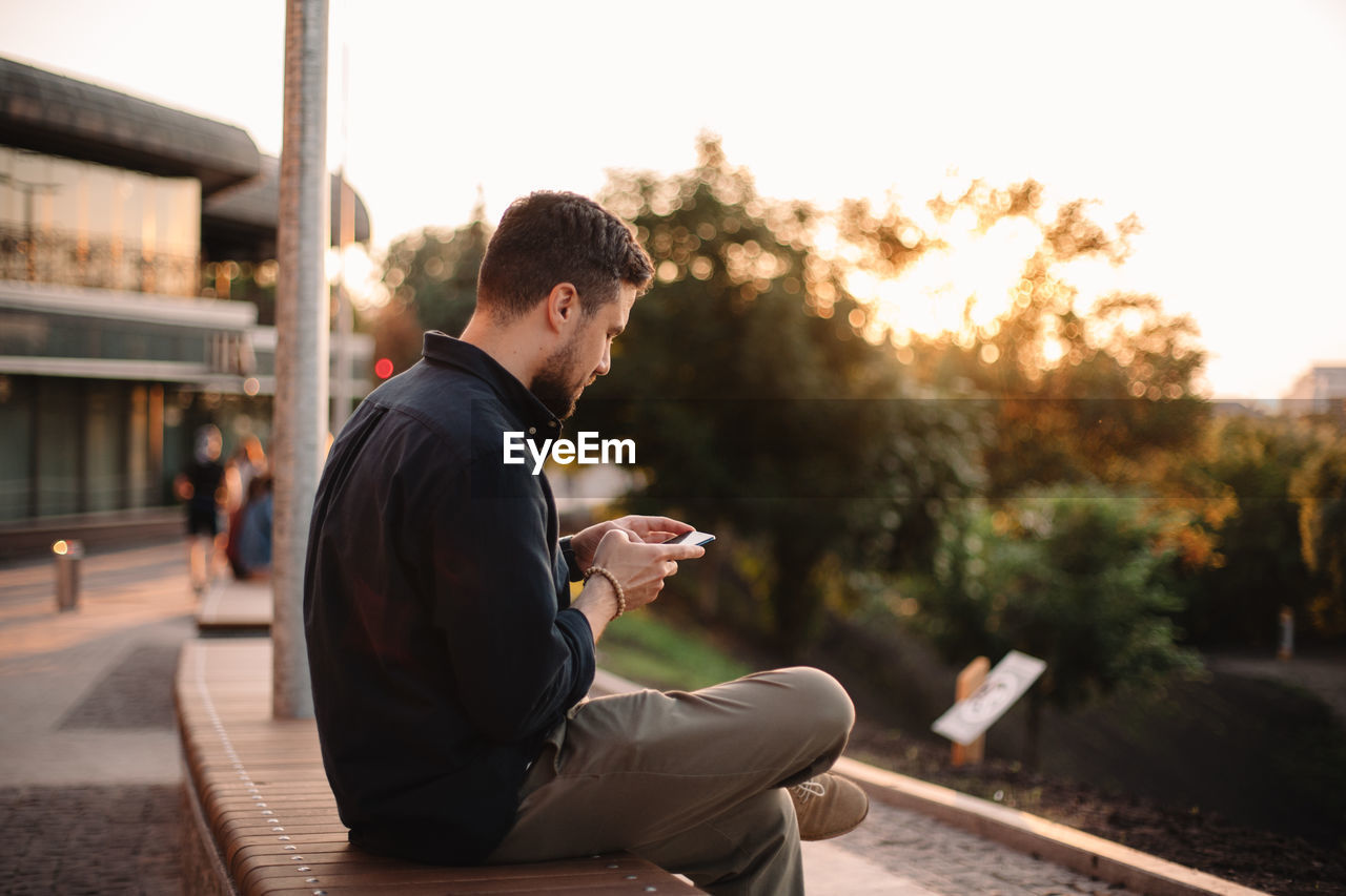 Happy man using smart phone sitting on bench at sunset