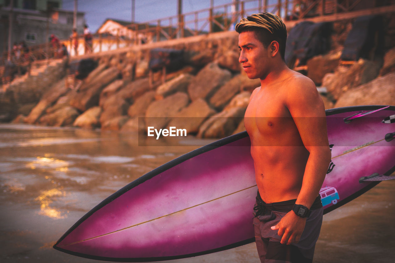 Young shirtless man carrying surfboard at beach