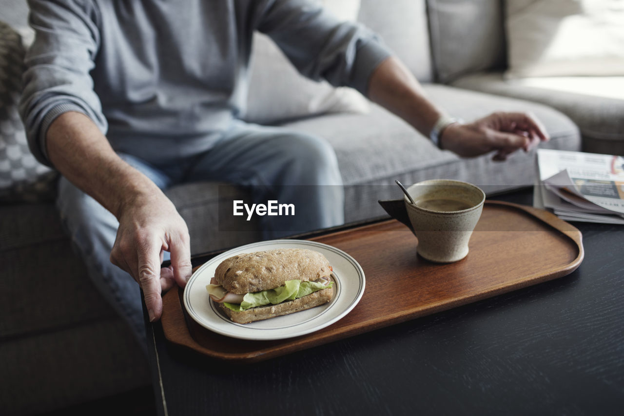 Midsection of senior man with sandwich and cup sitting on sofa at home