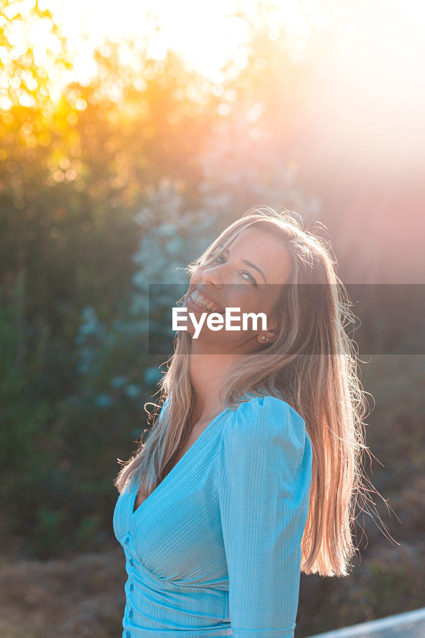 Portrait of smiling young woman standing in forest at sunset