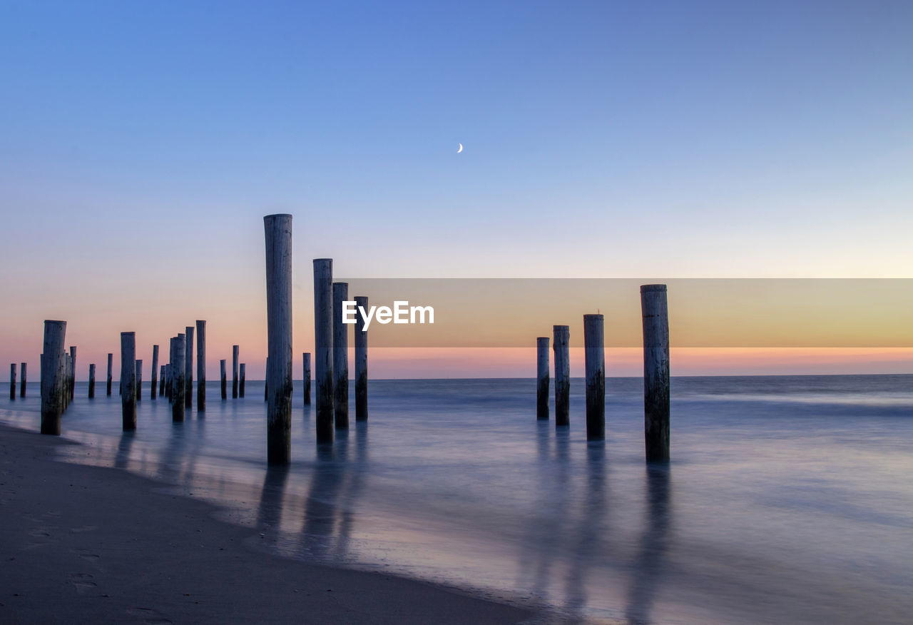 Wooden posts in sea against clear sky at sunset